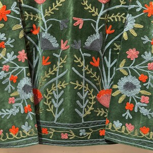 May include: Close-up of a green velvet garment with intricate floral embroidery. The embroidery features a variety of colors, including orange, pink, gray, and gold, creating a detailed and textured design. The floral pattern is repeated throughout the garment.