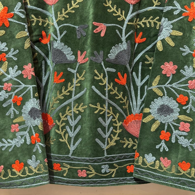May include: Close-up of a green velvet garment with intricate floral embroidery. The embroidery features a variety of colors, including orange, pink, gray, and gold, creating a detailed and textured design. The floral pattern is repeated throughout the garment.