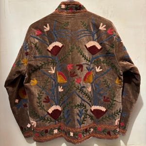 May include: A brown velvet jacket with detailed floral embroidery in red, pink, yellow, and blue. The jacket has a high collar and decorative stitching along the edges and sleeves, showcasing a bohemian style.
