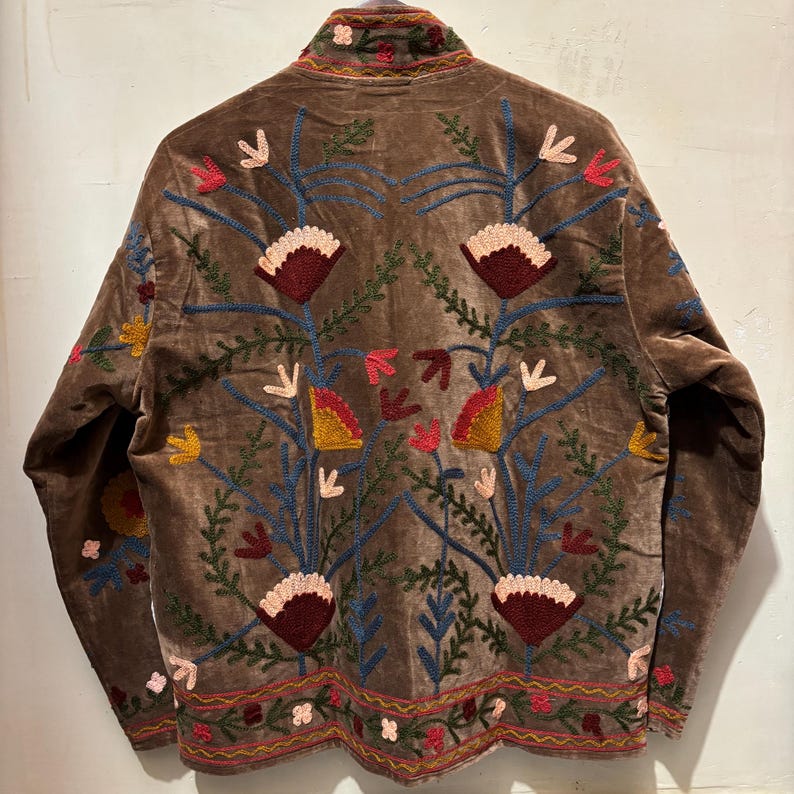 May include: A brown velvet jacket with detailed floral embroidery in red, pink, yellow, and blue. The jacket has a high collar and decorative stitching along the edges and sleeves, showcasing a bohemian style.