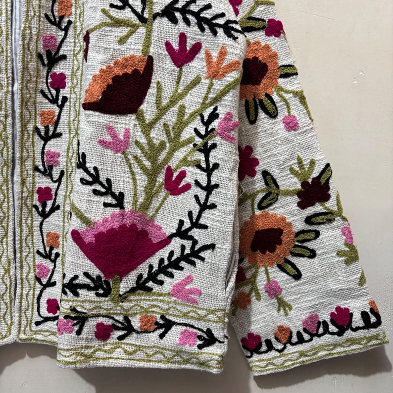 May include: A white jacket with intricate floral embroidery in shades of pink, orange, and burgundy. The jacket features a textured fabric and a detailed design, with black accents and green trim.