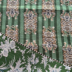 May include: A quilted textile featuring a repeating pattern of tigers on a green striped background. The reverse side displays a green fabric with a white and blue floral print. The two sides are joined by a thin, striped fabric edge binding.