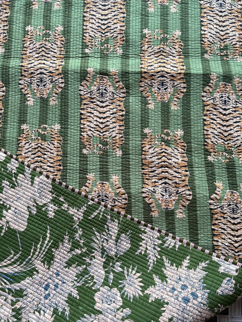 May include: A quilted textile featuring a repeating pattern of tigers on a green striped background. The reverse side displays a green fabric with a white and blue floral print. The two sides are joined by a thin, striped fabric edge binding.