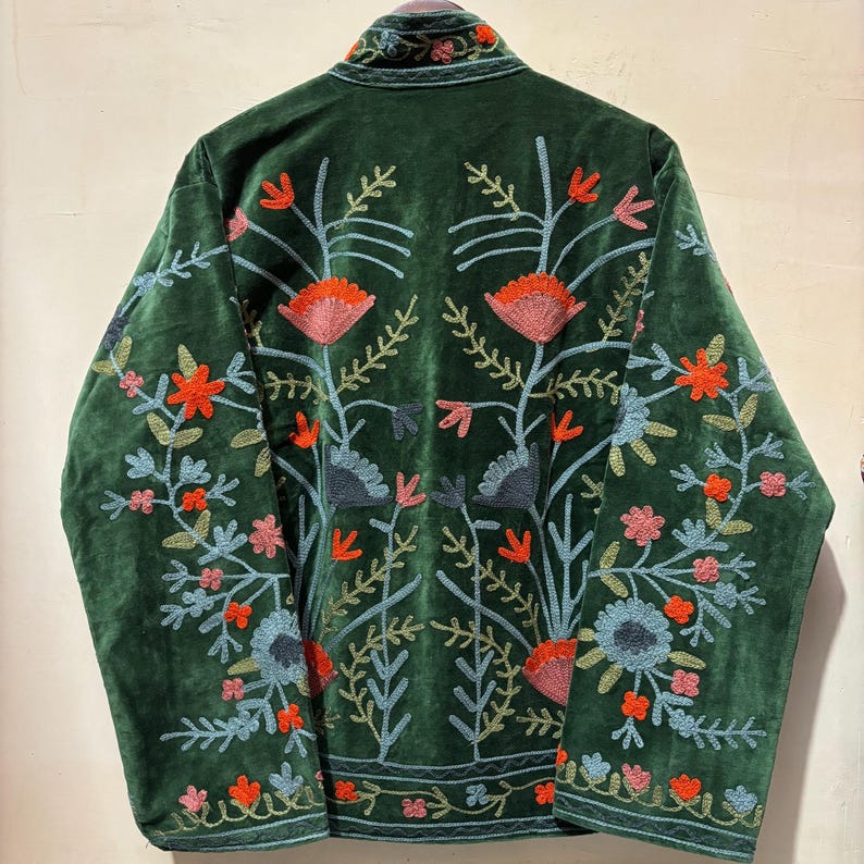 May include: A dark green velvet jacket with intricate floral embroidery in shades of orange, pink, and blue. The jacket features a high collar and detailed patterns on the back and sleeves, creating a bohemian aesthetic.