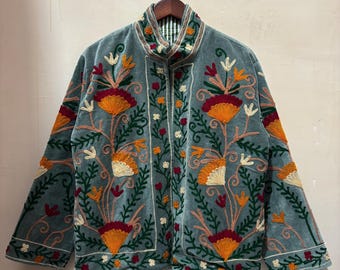 May include: A teal velvet jacket with intricate floral embroidery in orange, red, and cream. The jacket features a high collar and is displayed on a wooden hanger. The embroidery creates a striking contrast against the teal fabric.