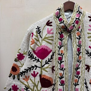 May include: Off-white jacket with colorful floral embroidery. The jacket features a collar and is adorned with pink, orange, and burgundy flowers, along with green leaves and stems. The jacket is hanging on a wooden hanger.