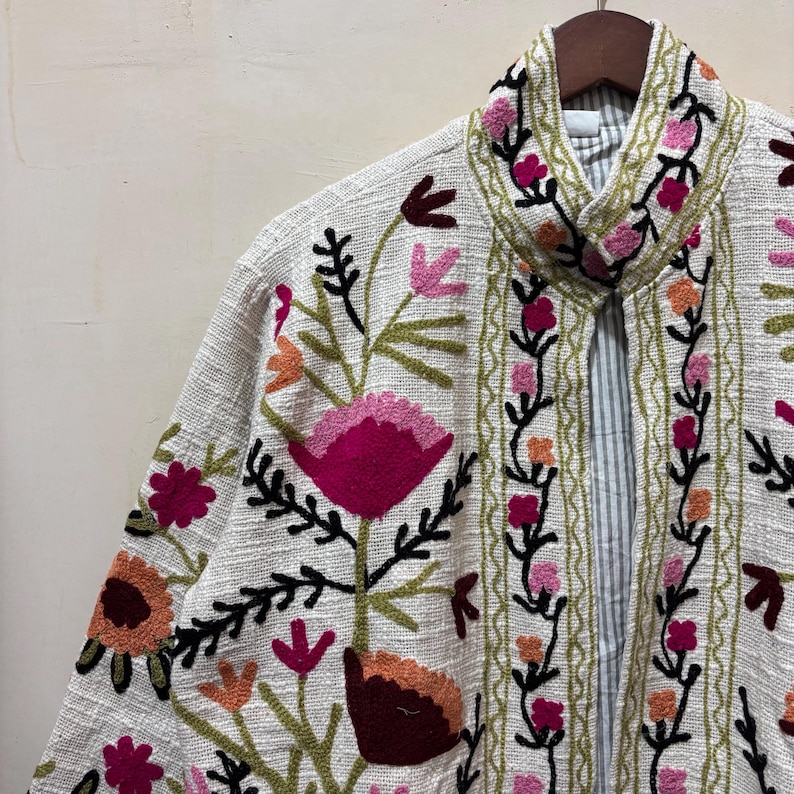 May include: Off-white jacket with colorful floral embroidery. The jacket features a collar and is adorned with pink, orange, and burgundy flowers, along with green leaves and stems. The jacket is hanging on a wooden hanger.