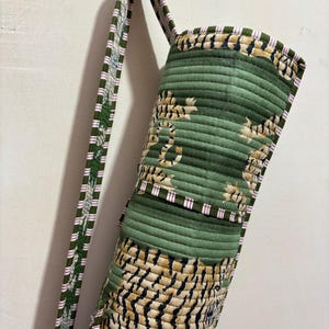 May include: A cylindrical quilted yoga mat bag featuring a green fabric base with a repeating tiger stripe pattern in black and tan. The bag includes a long shoulder strap with a green, white, and pink striped trim along the edges and seams.