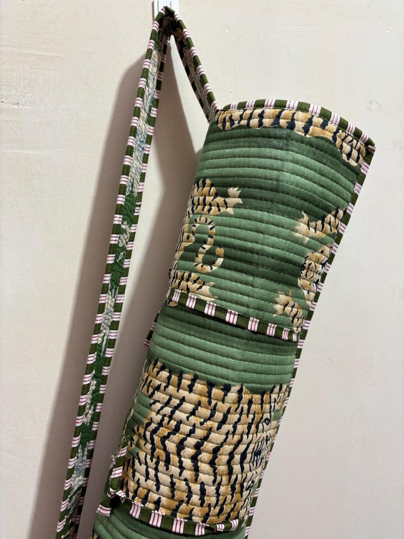May include: A cylindrical quilted yoga mat bag featuring a green fabric base with a repeating tiger stripe pattern in black and tan. The bag includes a long shoulder strap with a green, white, and pink striped trim along the edges and seams.
