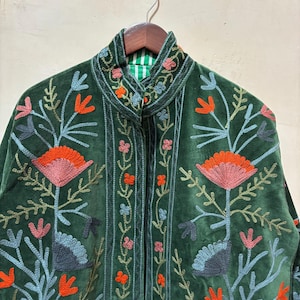 May include: A dark green velvet jacket with intricate floral embroidery in shades of orange, pink, blue, and gray. The jacket features a high collar and a striped green and white lining. The embroidery is detailed and covers the front and collar.
