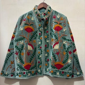 Può includere: Una giacca in velluto azzurro con intricati ricami floreali nei toni del rosso, arancione e crema. Il capo presenta un colletto alto, maniche lunghe e un'apertura frontale, con una fodera a righe verdi e bianche visibile sullo scollo.