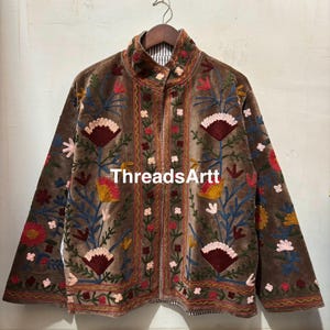 May include: Brown velvet jacket with vibrant floral embroidery. The jacket has a high collar and a full-length zip. The embroidery features red, yellow, and blue flowers. The text "ThreadsArtt" is visible on the jacket.