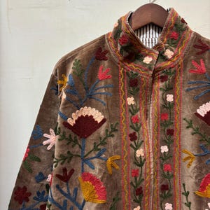 May include: Brown velvet jacket with colourful floral embroidery. The jacket has a high collar and a striped lining. The embroidery includes red, pink, yellow, and blue flowers and leaves, creating a vibrant design.