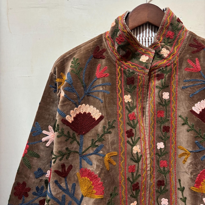 May include: Brown velvet jacket with colourful floral embroidery. The jacket has a high collar and a striped lining. The embroidery includes red, pink, yellow, and blue flowers and leaves, creating a vibrant design.