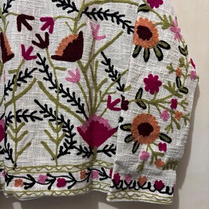 May include: Close-up of a white jacket with intricate floral embroidery. The design features flowers in shades of pink, orange, and burgundy, with green and black accents. The jacket's fabric has a textured appearance, and the embroidery is detailed.