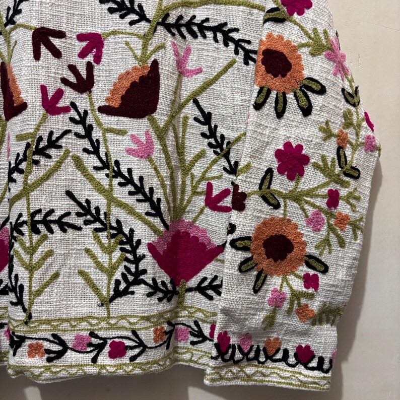 May include: Close-up of a white jacket with intricate floral embroidery. The design features flowers in shades of pink, orange, and burgundy, with green and black accents. The jacket's fabric has a textured appearance, and the embroidery is detailed.