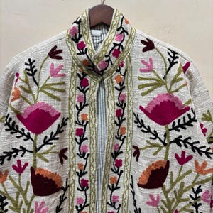 May include: Cream-colored jacket with intricate floral embroidery in shades of pink, orange, and green. The jacket features a high collar and a vertical floral design along the front edges. The embroidery creates a textured, artistic look.