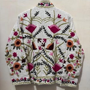 May include: A white jacket with colorful floral embroidery. The jacket features a detailed pattern of pink, orange, and green flowers and leaves. The embroidery covers the back and extends to the collar and hem, creating a vibrant, textured design.