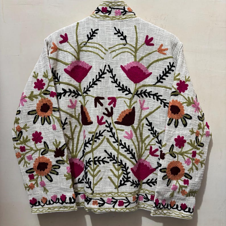 May include: A white jacket with colorful floral embroidery. The jacket features a detailed pattern of pink, orange, and green flowers and leaves. The embroidery covers the back and extends to the collar and hem, creating a vibrant, textured design.