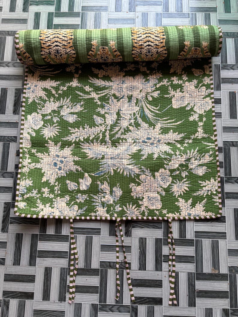 May include: A quilted fabric roll featuring a green background with a white floral pattern and a top section displaying a repeating tiger print. The edges are finished with a striped trim, and three sets of fabric ties hang from the bottom edge.