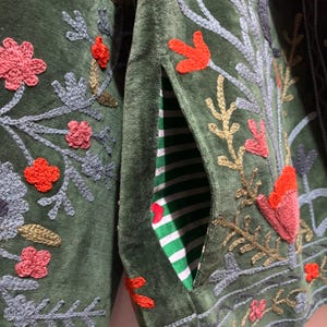 May include: Close-up of a dark green jacket with embroidered floral and leaf designs in shades of red, pink, gray, and gold. The jacket has a pocket with a green and white striped lining.