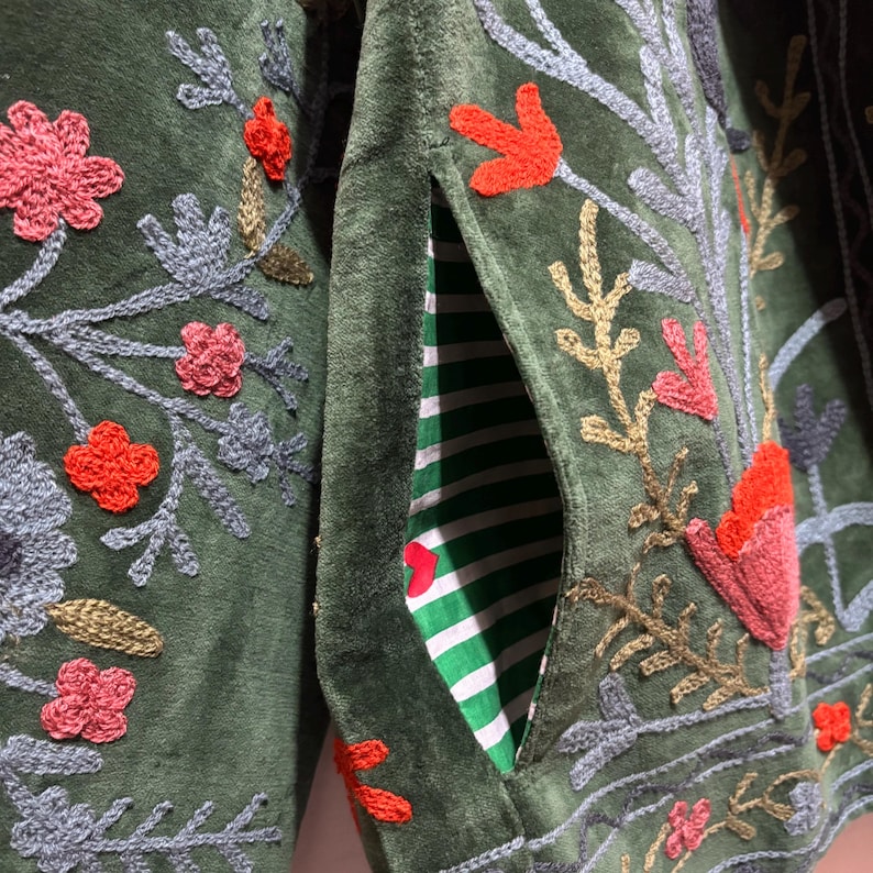 May include: Close-up of a dark green jacket with embroidered floral and leaf designs in shades of red, pink, gray, and gold. The jacket has a pocket with a green and white striped lining.