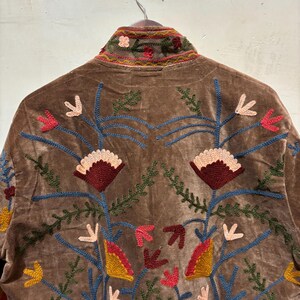 May include: Brown velvet jacket with vibrant floral embroidery. The jacket has a high collar and detailed designs in red, blue, yellow, and green. The embroidery features stylized flowers and leaf patterns.