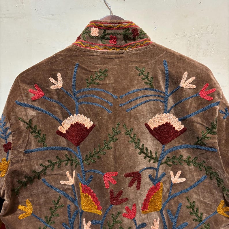 May include: Brown velvet jacket with vibrant floral embroidery. The jacket has a high collar and detailed designs in red, blue, yellow, and green. The embroidery features stylized flowers and leaf patterns.