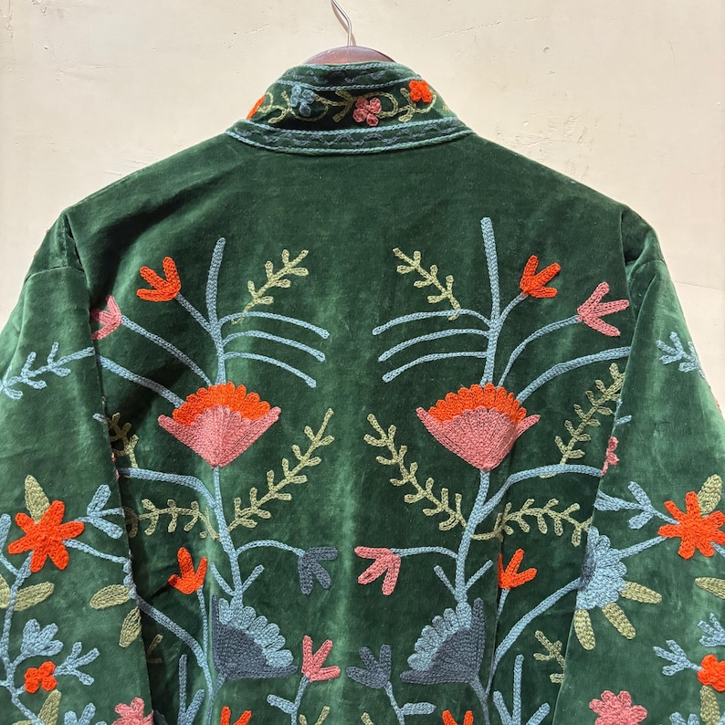 May include: A dark green velvet jacket with colorful floral embroidery. The embroidery features orange, pink, and blue flowers and leaves. The jacket is hanging on a wooden hanger.