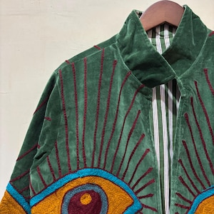 May include: Green velvet jacket with red embroidered lines and a colorful eye design. The jacket has a high collar and a striped lining. The design includes shades of teal, gold, and burgundy. The jacket is hanging on a wooden hanger.