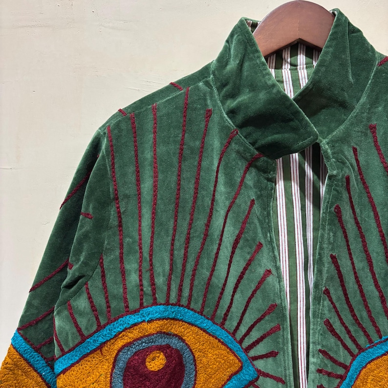 May include: Green velvet jacket with red embroidered lines and a colorful eye design. The jacket has a high collar and a striped lining. The design includes shades of teal, gold, and burgundy. The jacket is hanging on a wooden hanger.