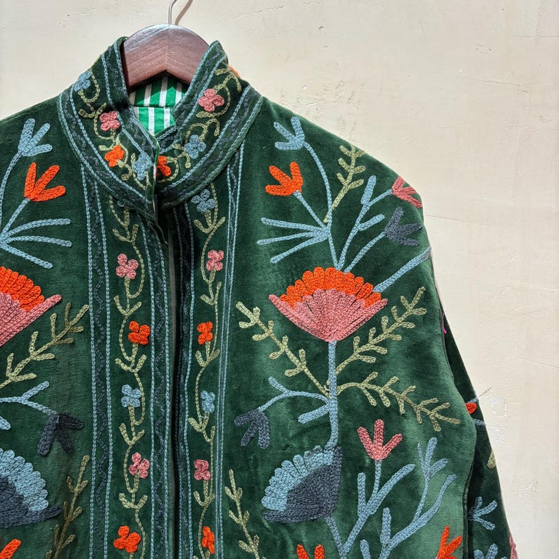 May include: A dark green velvet jacket with intricate floral embroidery in shades of orange, pink, blue, and green. The jacket features a high collar and a striped lining, showcasing a bohemian style.