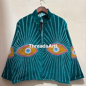 Op de afbeelding: Teal fluwelen jas met geborduurde oogontwerpen. De jas heeft een hoge kraag en een ritssluiting aan de voorkant. De ogen zijn geel met roze en blauwe accenten en de jas heeft verticale lijnen die van de ogen uitstralen. De tekst "ThreadsArtt" is zichtbaar.