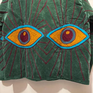 May include: Green velvet jacket with embroidered eye design. The jacket features two large, stylized eyes in shades of yellow, blue, and burgundy. The eyes are surrounded by radiating lines in burgundy. The jacket is a unique piece of wearable art.