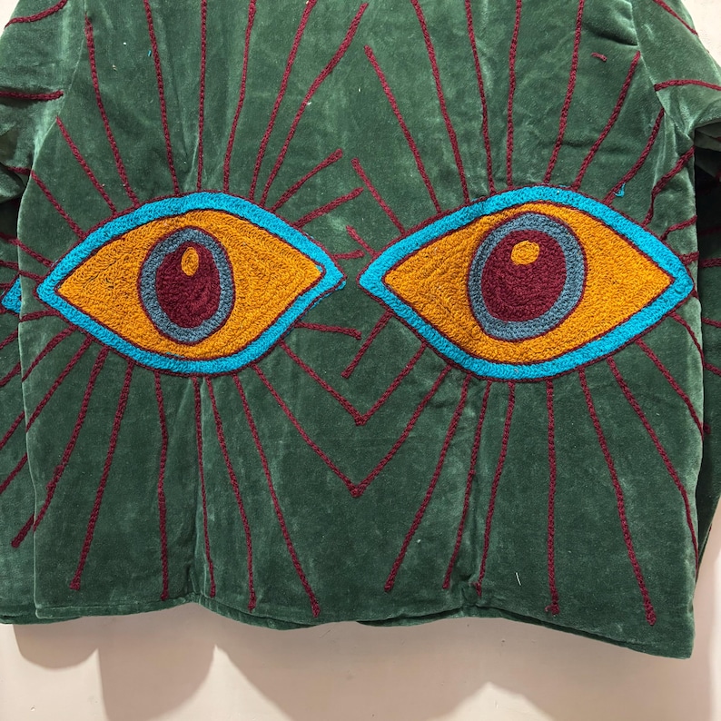 May include: Green velvet jacket with embroidered eye design. The jacket features two large, stylized eyes in shades of yellow, blue, and burgundy. The eyes are surrounded by radiating lines in burgundy. The jacket is a unique piece of wearable art.