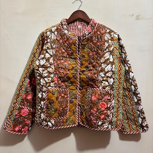 Op de afbeelding: Een gewatteerd jack met een patchworkontwerp in bruine, mosterdgele en witte bloemenprints, geaccentueerd door verticale zigzagstrepen in petrol, rood en geel. Het jack heeft een ronde halslijn, lange mouwen, twee voorzakken en een knoopsluiting.