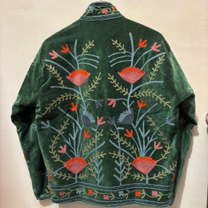 May include: A dark green velvet jacket with intricate floral embroidery in shades of orange, pink, and blue. The embroidered design covers the back and collar of the jacket, creating a striking visual contrast. The jacket has a relaxed fit and a classic silhouette.
