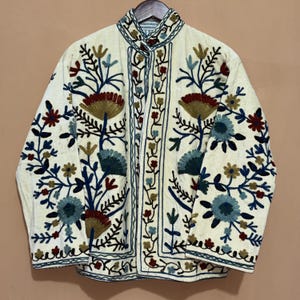 May include: Cream-colored jacket with intricate floral embroidery in shades of blue, red, and gold. The jacket features a high collar and button closure. The embroidery creates a detailed pattern across the front and sleeves, showcasing a bohemian style.