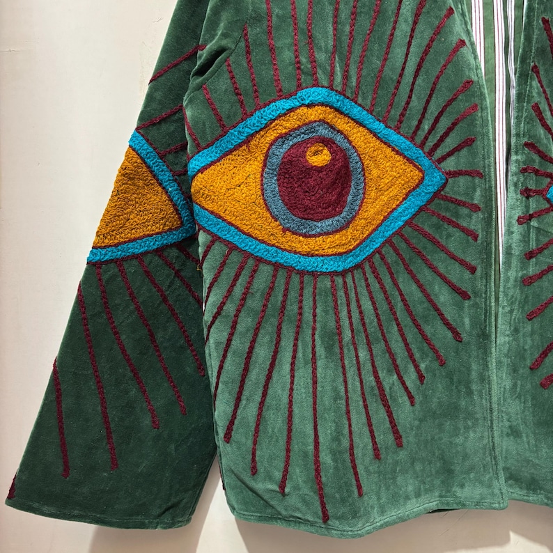 May include: Green velvet jacket with embroidered eye design. The jacket features a large, stylized eye in shades of yellow, blue, and burgundy, with radiating lines in burgundy. The sleeve has a triangular design in yellow and blue.