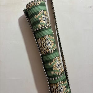 May include: A cylindrical quilted yoga mat bag in a sage green color featuring a repeating pattern of tigers with black stripes. The bag includes a long, adjustable shoulder strap with a dark green and white striped trim along the edges.