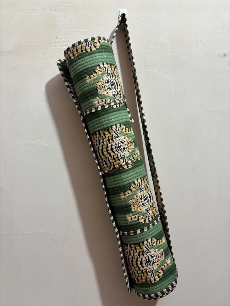 May include: A cylindrical quilted yoga mat bag in a sage green color featuring a repeating pattern of tigers with black stripes. The bag includes a long, adjustable shoulder strap with a dark green and white striped trim along the edges.