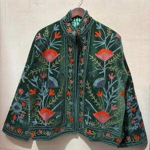 May include: A dark green velvet jacket with intricate floral embroidery in shades of orange, blue, and pink. The jacket features a high collar and a striped green and white lining. The embroidery creates a botanical design across the jacket's surface.