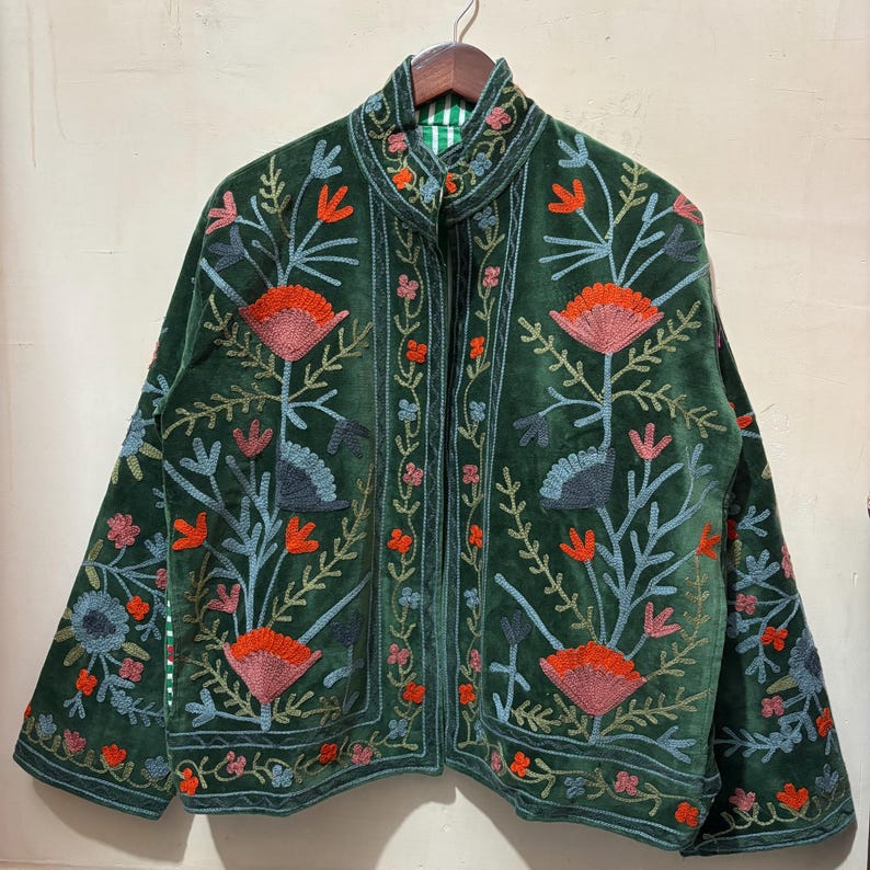 May include: A dark green velvet jacket with intricate floral embroidery in shades of orange, blue, and pink. The jacket features a high collar and a striped green and white lining. The embroidery creates a botanical design across the jacket's surface.