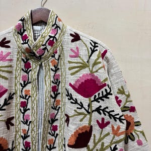 May include: A cream-colored jacket with intricate floral embroidery in shades of pink, orange, and green. The jacket features a high collar and a textured fabric, showcasing a bohemian style.