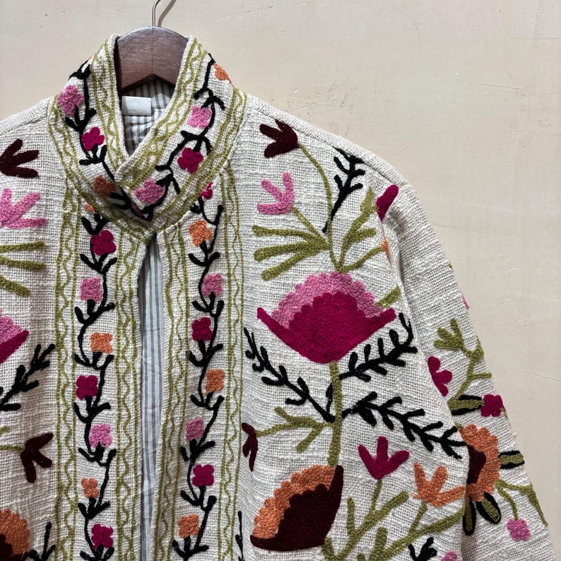 May include: A cream-colored jacket with intricate floral embroidery in shades of pink, orange, and green. The jacket features a high collar and a textured fabric, showcasing a bohemian style.
