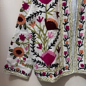 May include: White jacket with colorful floral embroidery. The jacket features a variety of flowers in shades of pink, orange, and burgundy, with green leaves and black stems. The embroidery is detailed and covers the front and sleeves.