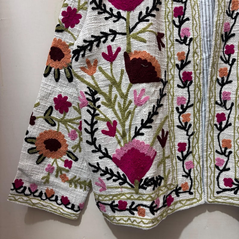 May include: White jacket with colorful floral embroidery. The jacket features a variety of flowers in shades of pink, orange, and burgundy, with green leaves and black stems. The embroidery is detailed and covers the front and sleeves.