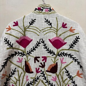 May include: A white jacket with intricate floral embroidery. The design features large magenta flowers, smaller pink and orange blossoms, and black and green foliage. The jacket is hanging on a hanger.