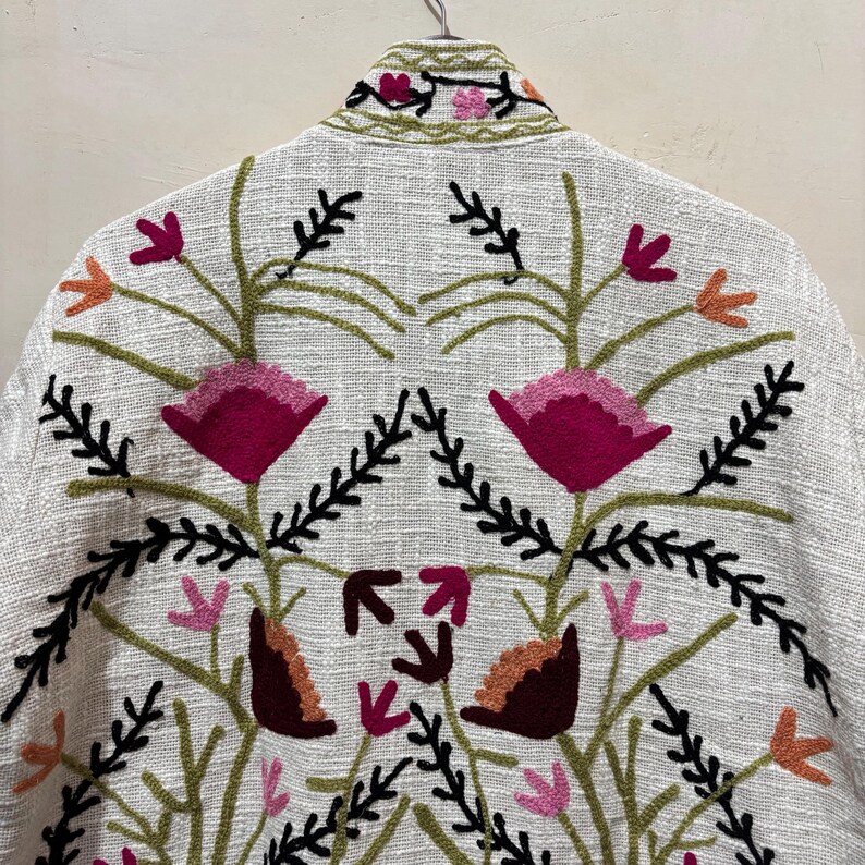 May include: A white jacket with intricate floral embroidery. The design features large magenta flowers, smaller pink and orange blossoms, and black and green foliage. The jacket is hanging on a hanger.