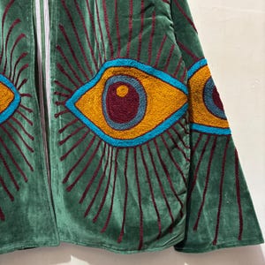 May include: Green velvet jacket with embroidered eye design. The jacket features large, stylized eyes in shades of yellow, teal, and burgundy. Vertical burgundy lines radiate from the eyes. The jacket has a relaxed fit and a soft texture.